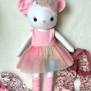 May include: A white plush mouse doll with large pink ears, wearing a pink dress with a rainbow tulle skirt. Additional outfits with floral patterns and a pink headband are included.
