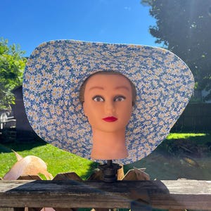 May include: A wide-brimmed sun hat featuring a vibrant blue fabric base covered in a dense pattern of small white daisies with yellow centers. The hat is displayed on a mannequin head, showcasing the full circular brim and the floral print design.