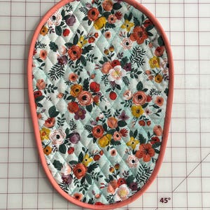 Puede incluir: Salvamanteles acolchado de forma ovalada con un estampado floral. La tela presenta un fondo azul claro con un patrón repetido de flores naranjas, amarillas y rojas. El borde está ribeteado con un ribete color coral. Mide unos 20 cm de largo.