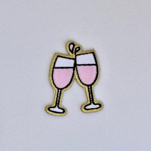 Puede incluir: Un parche bordado en oro con dos copas de vino rosa brindando.