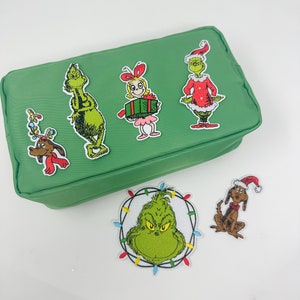 May include: A green zippered pouch with five embroidered patches featuring characters from the Grinch movie. The patches include the Grinch, Cindy Lou Who, Max the dog, and two different images of the Grinch.