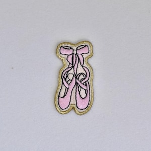 May include: Pink ballet shoes with a gold outline and a pink bow. The shoes are embroidered on a white fabric.