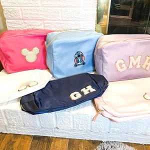 May include: Six different cosmetic bags with various embroidered designs. The bags are pink, light blue, lavender, white, and navy blue. The designs include Mickey Mouse, Stitch, the letters "GMH", the letters "GH", and a simple Mickey Mouse head.