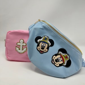 以下が含まれることがあります： 2つのファニーパック。1つはピンクで白い錨と金のアクセントが付いており、もう1つは薄いブルーで、帽子をかぶったミッキーマウスの刺繍が2つ施されています。