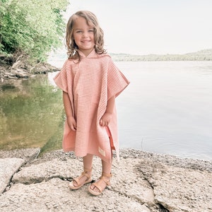May include: A young girl wearing a pink hooded towel, standing on a rocky surface by a body of water.