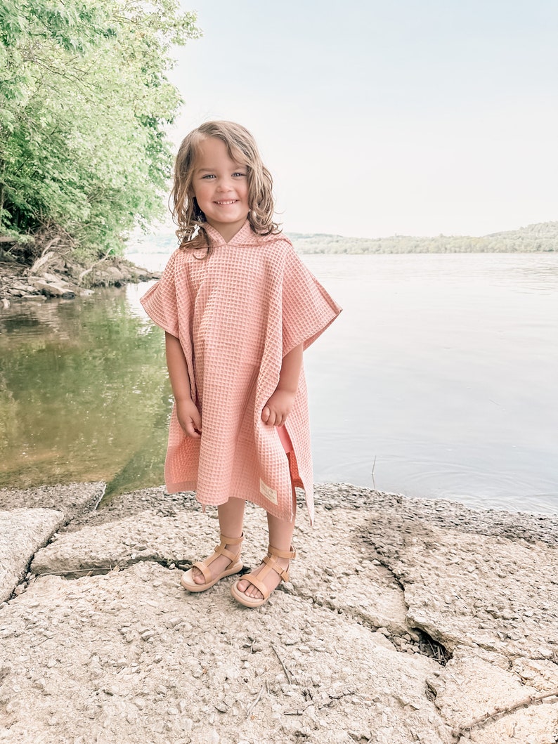 May include: A young girl wearing a pink hooded towel, standing on a rocky surface by a body of water.
