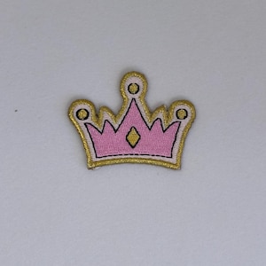 May include: A pink and gold embroidered crown patch. The crown has a pointed top and five points on each side. The crown is outlined in gold thread and has a diamond shape in the center.