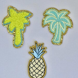 May include: Three embroidered iron-on patches with a tropical theme. A yellow palm tree, a blue palm tree, and a white pineapple with a blue top.