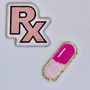 May include: Two embroidered patches: one in the shape of the Rx symbol in pink and white with a silver border, and a pink and white capsule-shaped patch with a gold glitter border. The patches are on a white background.