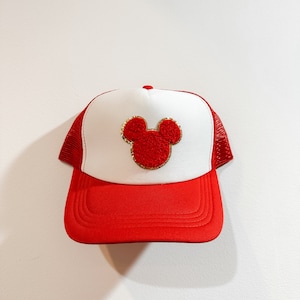 May include: A red and white trucker hat with a red Mickey Mouse head embroidered on the front. The Mickey Mouse head has gold stitching around the outline.