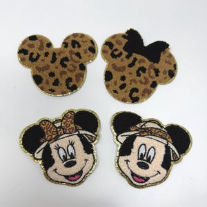 Może przedstawiać: Cztery haftowane naszywki z motywami Myszki Miki i Myszki Minnie. Dwie naszywki mają uszy w panterkę, a pozostałe dwie uśmiechnięte twarze z kapeluszami i kokardami. Wszystkie naszywki mają złote, błyszczące obramowania.