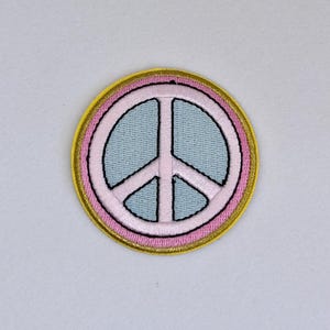 May include: Embroidered peace sign patch with a light blue center, pink outline, and gold border. The peace symbol is a black line drawing. This patch can be ironed on or sewn onto clothing or accessories.