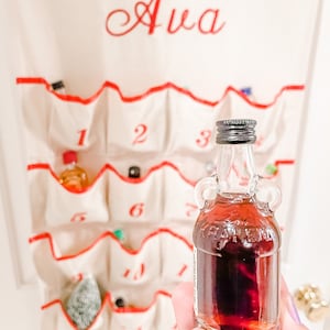May include: A small glass bottle with a red liquid inside. The bottle has a silver cap and a label that says "Frankenmuth". The bottle is in front of a white canvas advent calendar with the word "Ava" embroidered on it.