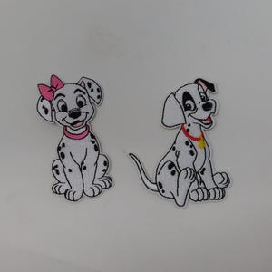 Könnte beinhalten: Zwei aufgestickte Aufnäher mit Dalmatiner-Welpen. Einer trägt eine rosa Schleife und Halsband, der andere ein rotes Halsband mit gelbem Anhänger. Beide sind weiß mit schwarzen Flecken, vor weißem Hintergrund.