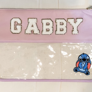 May include: A light purple zippered pouch with a clear plastic window. The pouch has the name "GABBY" in white letters with gold glitter on the front. A blue cartoon character with red accents is visible through the window.