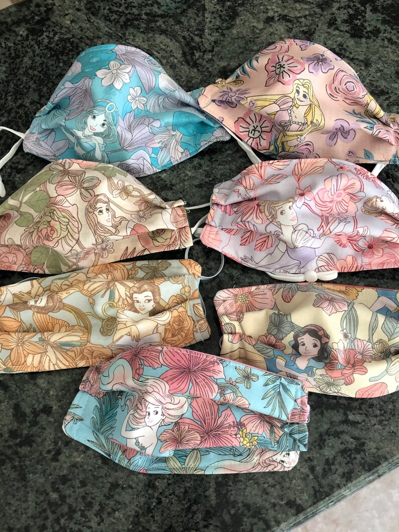 May include: Set of six colorful fabric face masks featuring different Disney princesses. Each mask has a floral pattern and a different princess image. The princesses include Ariel, Belle, Cinderella, Snow White, Rapunzel, and Jasmine.