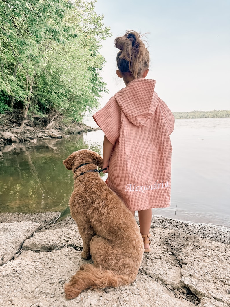 May include: A young girl wearing a pink waffle-knit hooded towel with the name "Alexandria" embroidered on the back, stands by a body of water with a brown dog sitting beside her.