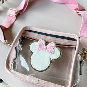 May include: A clear plastic crossbody bag with a pink strap and a pink and white Minnie Mouse applique. The bag has a zipper closure and a gold chain.