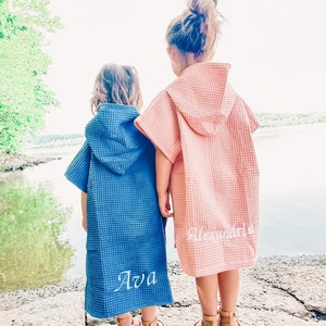 May include: Two children wearing hooded towels, one blue and one pink, with the names "Ava" and "Alexandria" embroidered on the back. The children are standing on a rocky surface by a lake.