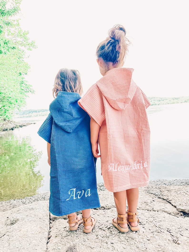 May include: Two children wearing hooded towels, one blue and one pink, with the names "Ava" and "Alexandria" embroidered on the back. The children are standing on a rocky surface by a lake.
