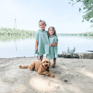 May include: Two young girls wearing light blue hooded towels stand on a sandy beach with a brown dog lying in front of them. The girls are smiling and hugging each other. The dog is looking at the camera.