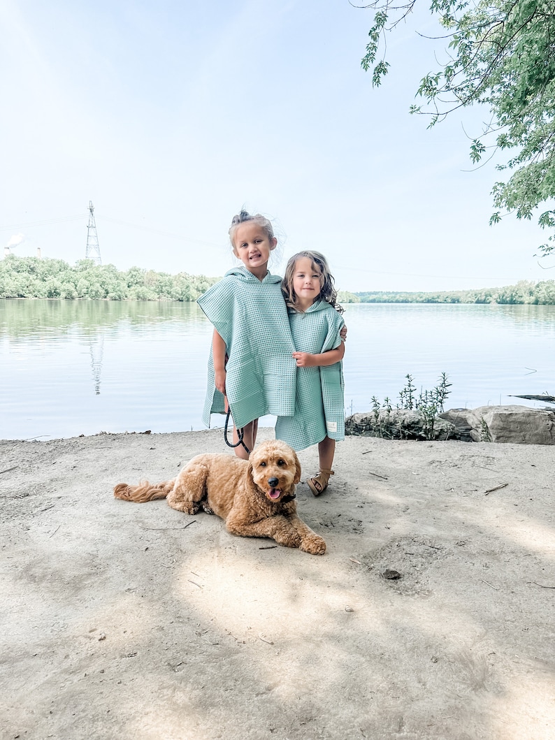 May include: Two young girls wearing light blue hooded towels stand on a sandy beach with a brown dog lying in front of them. The girls are smiling and hugging each other. The dog is looking at the camera.
