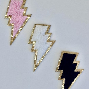 Könnte beinhalten: Drei dekorative Blitz-Patches in Rosa, Weiß und Marineblau. Jeder Patch hat einen goldglitzernden Rand. Die rosa und weißen Blitze haben ein strukturiertes, flauschiges Aussehen.