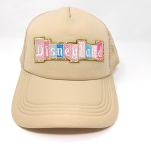 May include: Tan trucker hat with a curved brim and mesh back. The front features the word "Disneyland" in colorful, embroidered letters. The hat is a neutral color, suitable for various outfits and occasions.