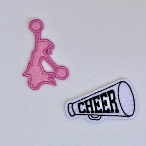 May include: Two iron-on patches for clothing. One is pink and depicts a cheerleader holding pom-poms. The other is white and depicts a megaphone with the word "CHEER" in black.