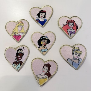 Op de afbeelding: Zeven geborduurde strijkpatches in de vorm van harten met verschillende Disney-prinsessen. Elke patch heeft een roze achtergrond met een gouden glitterrand en een andere prinses in het midden. De prinsessen zijn Sneeuwwitje, Assepoester, Ariel, Belle, Jasmine, Tiana en Rapunzel.