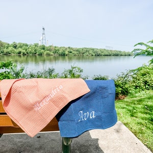 May include: Two waffle weave towels, one pink and one blue, are folded and placed on a wooden bench. The towels are personalized with the names "Alexandria" and "Ava" in white embroidery.