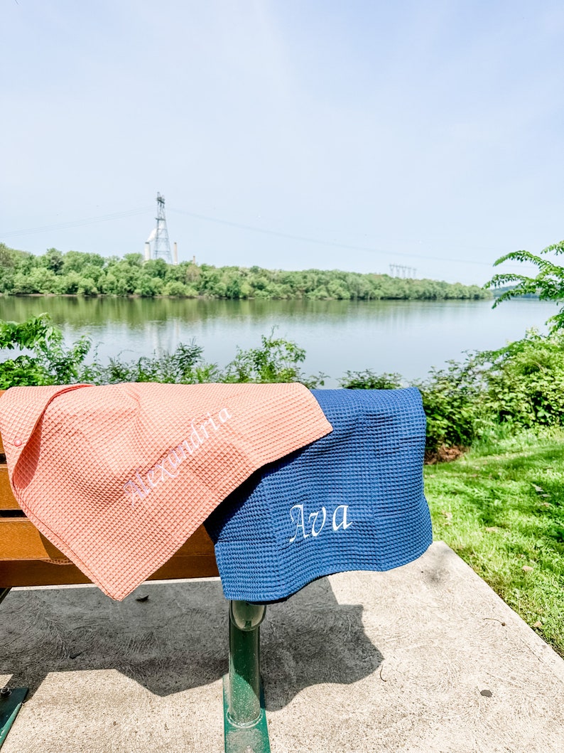 May include: Two waffle weave towels, one pink and one blue, are folded and placed on a wooden bench. The towels are personalized with the names "Alexandria" and "Ava" in white embroidery.