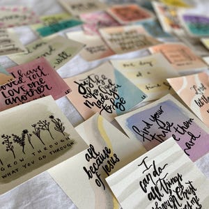 以下が含まれることがあります： パステルカラーの水彩画風の背景に、黒の手書き文字でインスピレーションを与える引用句が書かれた正方形のカードのコレクション。「Grow through what you go through」や「I can do all things through Christ」といったユニークなフレーズが各カードに記されています。