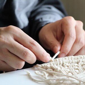May include: Close-up of a person's hands working on a macrame project. The person is using white cord to create a knot pattern. The macrame is on a white surface.