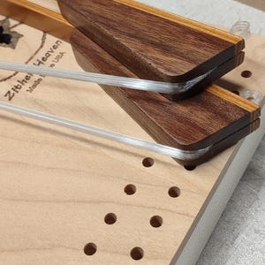 May include: A wooden zither instrument with a light-colored body and dark brown wooden bridges. The zither has clear strings and decorative leaf and vine inlays. The text "Zither Heaven" is visible on the instrument.