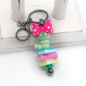 May include: A whimsical keychain featuring a green worm with glasses, a pink bow with white polka dots, and a stack of colorful books. The keychain has a silver-tone clasp and a small, decorative bead.