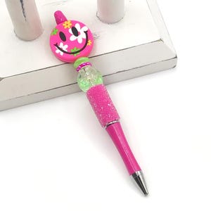 May include: A pink pen with a sparkly pink and green glitter design. The top of the pen is a round, pink smiley face with white flowers and green leaves.