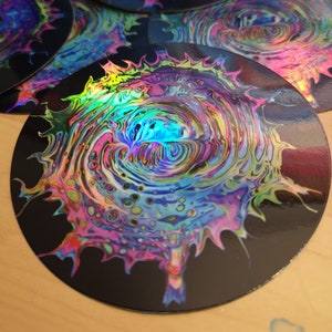 May include: A round sticker with a swirling, abstract design in shades of blue, green, pink, and purple. The design is reminiscent of a galaxy or nebula. The sticker has a glossy finish and a black background.