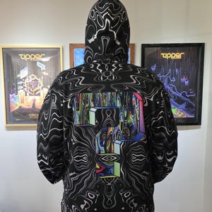 Puede incluir: Una sudadera con capucha negra que presenta un patrón repetitivo de líneas onduladas blancas en toda la prenda. La parte posterior muestra un gráfico grande y central de un signo de interrogación con un diseño líquido multicolor.