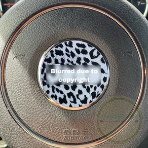 May include: A black and white leopard print steering wheel cover with a gold trim. The cover is fitted to a black leather steering wheel with a silver "SRS AIRBAG" label.