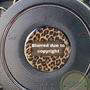 May include: A black leather steering wheel with a brown and black leopard print design. The steering wheel has a silver trim and a silver horn button. The text "Blurred due to copyright" is visible in the centre of the steering wheel.