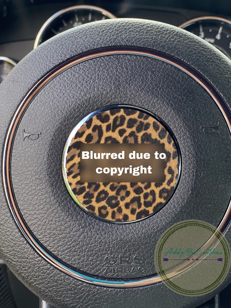 May include: A black leather steering wheel with a brown and black leopard print design. The steering wheel has a silver trim and a silver horn button. The text "Blurred due to copyright" is visible in the centre of the steering wheel.