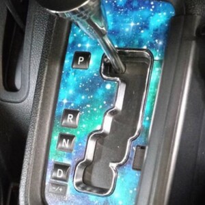 May include: A blue and green galaxy-themed car gear shift plate with the letters "P", "R", "N", "D", and "+/-" printed on it.
