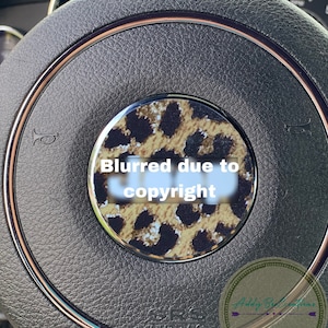 May include: A close-up of a black leather steering wheel with a gold trim. The centre of the steering wheel has a round, leopard print sticker with the text "Blurred due to copyright".