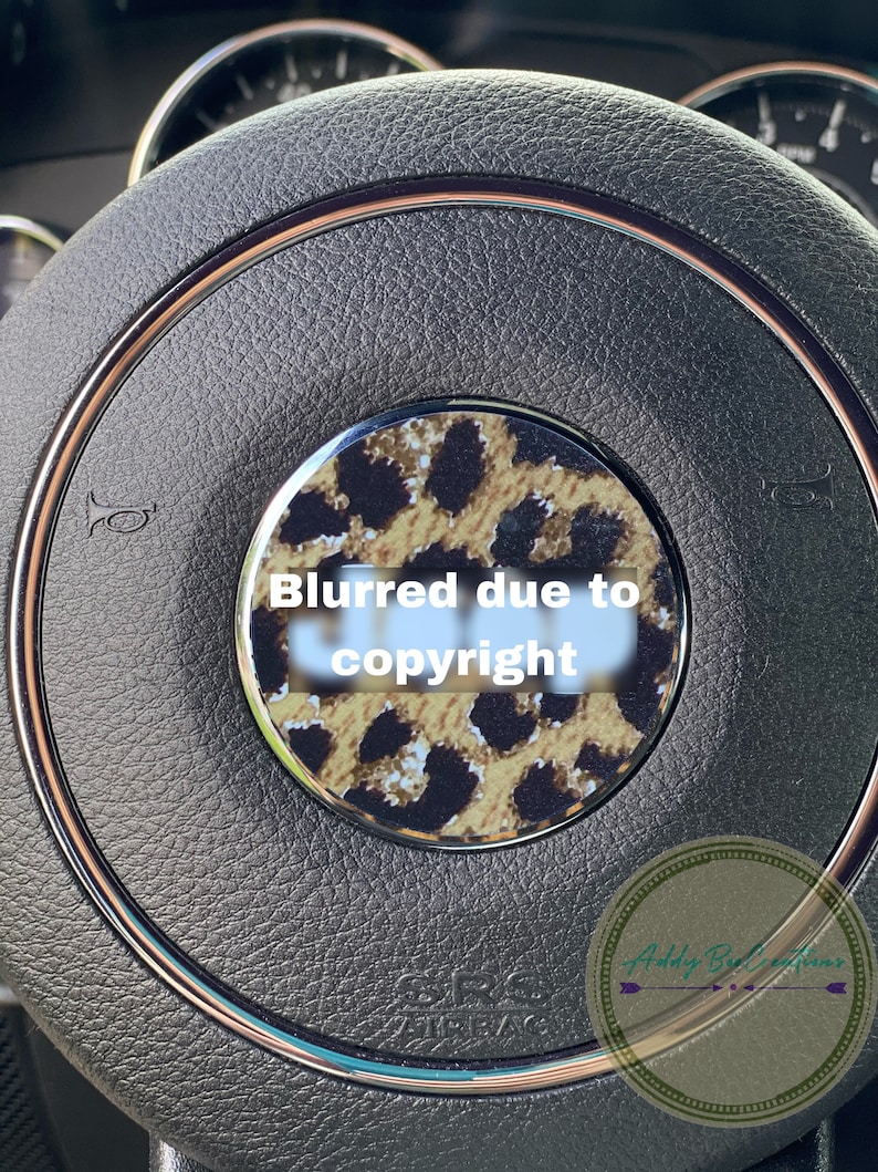 May include: A close-up of a black leather steering wheel with a gold trim. The centre of the steering wheel has a round, leopard print sticker with the text "Blurred due to copyright".