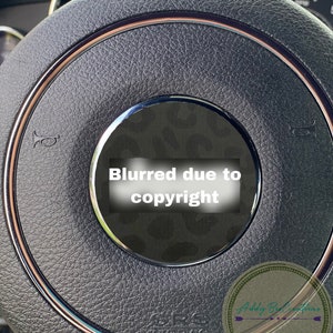 May include: Close-up of a black leather steering wheel with a brown trim. The steering wheel has a silver horn button and a black and white patterned centre cap. The text "Blurred due to copyright" is visible on the centre cap.