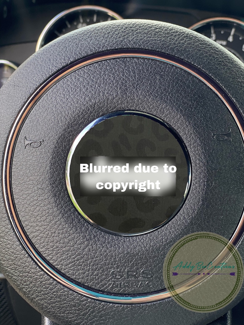 May include: Close-up of a black leather steering wheel with a brown trim. The steering wheel has a silver horn button and a black and white patterned centre cap. The text "Blurred due to copyright" is visible on the centre cap.