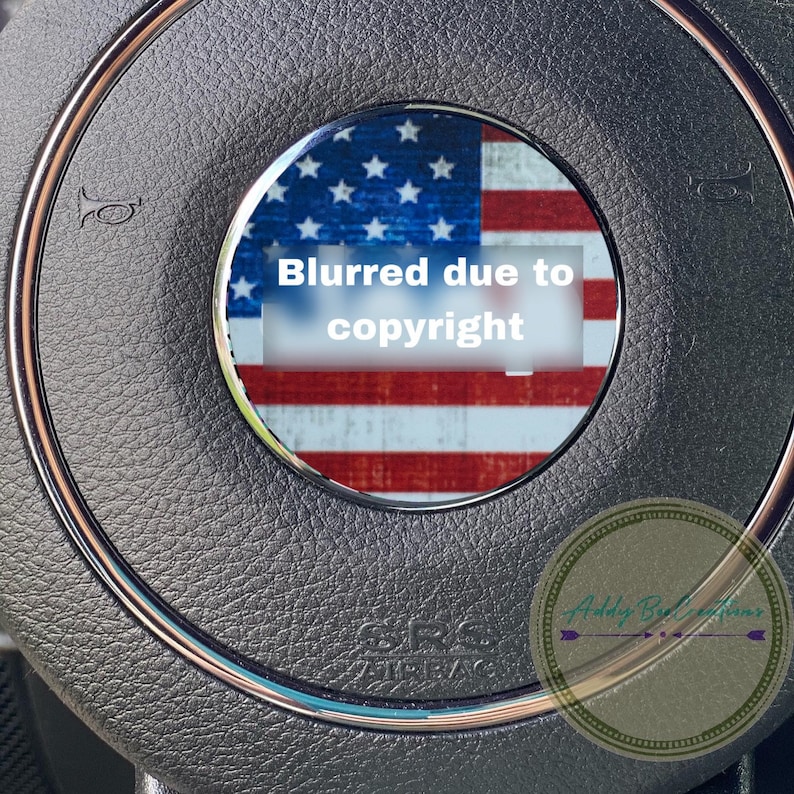 May include: A close-up of a black steering wheel with a silver trim. The center of the steering wheel has a round, silver-rimmed circle with an American flag inside. The flag is blurred due to copyright. The text "SRS AIRBAG" is visible below the flag. The text "Blurred due to copyright" is visible on the flag.