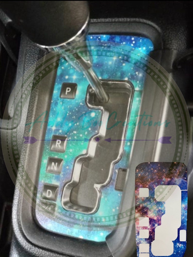 May include: A galaxy-themed car gear shift cover with a blue, green, and purple colour scheme. The cover features a galaxy print with stars and nebulae. The cover is designed to fit over the gear shift lever and has cutouts for the gear shift buttons. The cover also includes a matching galaxy-themed key fob cover.