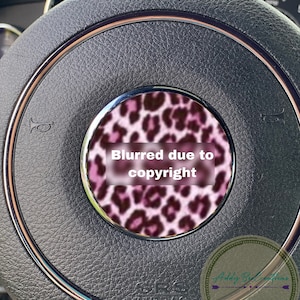 May include: A close-up of a black leather steering wheel with a pink and black leopard print design in the centre. The text "Blurred due to copyright" is visible in the centre of the design.